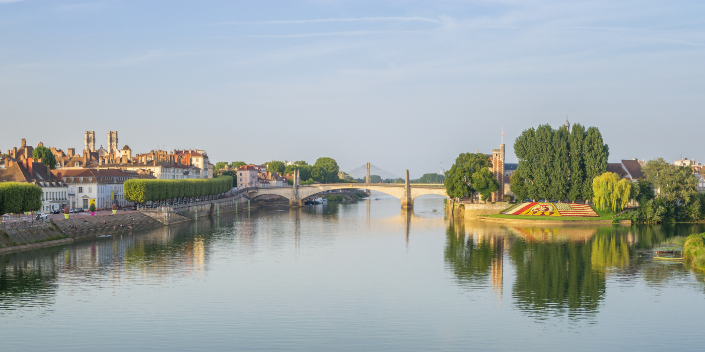 Das Bild zeigt eine malerische Szene entlang des Flusses Rhône in einer französischen Stadt. Auf der linken Seite des Bildes sind historische Gebäude mit roten Ziegeldächern zu sehen, während auf der rechten Seite ein bunter Blumenmuster, möglicherweise ein Gartendesign, zu erkennen ist. Ein Brücke verbindet beide Ufer, und der ruhige Fluss spiegelt die Umgebung wider. Der Himmel ist klar, und das Bild vermittelt eine friedliche, frühe Morgenstimmung.