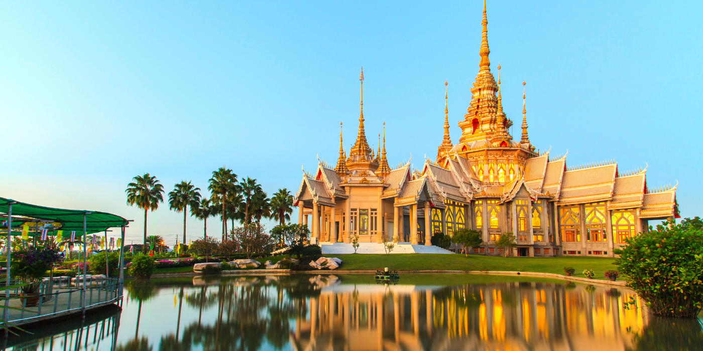 Das Bild zeigt ein wunderschönes asiatisches Tempelgebäude, das in goldenen und grauen Farbtönen erleuchtet ist. Es steht inmitten eines gepflegten Gartens, der von Palmen und grünen Pflanzen umgeben ist. Der Tempel reflektiert auf der Oberfläche eines ruhigen Teiches, was die friedliche und majestätische Atmosphäre verstärkt. Der Himmel ist klar und der Sonnenaufgang oder -untergang sorgt für sanfte Beleuchtung und eine warme Farbpalette.