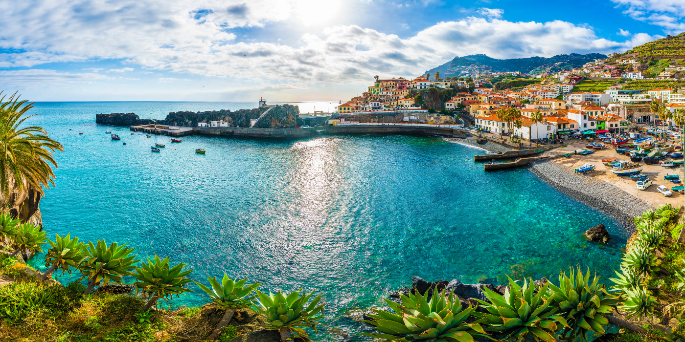 Das Bild zeigt eine malerische Küstenlandschaft, wahrscheinlich von der Insel Madeira. Im Vordergrund sind grüne Agavenpflanzen sichtbar, die entlang der Küste wachsen, während sich der klare, türkisfarbene Ozean erstreckt. Auf dem Wasser schwimmen verschiedene kleine Boote, und an der Küste ist ein malerisches Fischerdorf zu sehen, mit bunten Häusern und einer steinigen Strandpromenade. Die Häuser sind in hellen Tönen gehalten, und es gibt mehrere Palmen, die den tropischen Charme der Gegend verstärken. Im Hintergrund sieht man die grünen Hügel der Insel, die mit Wohnhäusern und terrassenförmigen Feldern bedeckt sind. Die Szene wird von einem strahlend blauen Himmel und sanften Wolken begleitet.